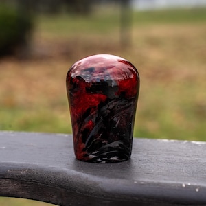 Może przedstawiać: Ręcznie wykonana, błyszcząca gałka zmiany biegów o unikalnym wzornictwie. Góra jest głęboko czerwona, przechodząca w czerń u podstawy. Gałka wykonana jest z wytrzymałego materiału i jest przeznaczona do dźwigni zmiany biegów w samochodzie.