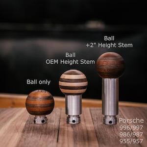 Op de afbeelding: Drie houten, kogelvormige auto-accessoires met metalen stelen. De ballen hebben verschillende houtnerfpatronen. Tekst op de afbeelding luidt "Ball only", "Ball OEM Height Stem" en "Ball +2" Height Stem". De tekst "Porsche 996/997 986/987 955/957" is ook zichtbaar.