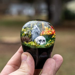 Op de afbeelding: Een helder, bolvormig hars object met een miniatuur kerkhofscène. De scène bevat een schedel, een grafsteen en kleurrijk gebladerte. De basis van het object is zwart.