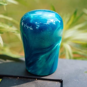 May include: A handcrafted, glossy, teal and blue resin object. The object has a rounded top and a slightly flared base. The colours blend and swirl, creating a marbled effect. The object is sitting on a dark surface, with a blurred green background.