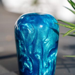 Könnte beinhalten: Zylindrisches Objekt mit einem glänzenden, wirbelnden blau-türkisen Design. Das Objekt hat eine abgerundete Oberseite und eine dunkle Basis. Die Oberfläche hat einen schimmernden, glitzernden Effekt, der eine dynamische visuelle Textur erzeugt. Der Hintergrund ist verschwommen, mit Andeutungen von grünem Laub.