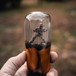 May include: A clear glass vial with a miniature figure inside, set in a dark soil base. The vial is encased in a wooden base with orange and black marbling. The figure appears to be a zombie.
