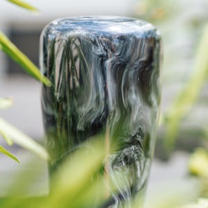 May include: A cylindrical object with a swirling pattern of black, white, and gray. The object has a rounded top and is set against a blurred background of green foliage. The surface appears glossy.