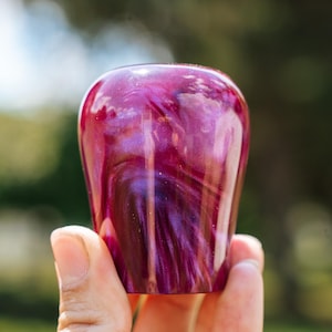 May include: A handcrafted, decorative bottle stopper with a swirling, marbled design in shades of deep magenta and purple. The stopper has a rounded top and a slightly tapered shape. The image is set against a blurred green background.