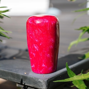 May include: A vibrant red, cylindrical object with a glossy finish. The surface has subtle pink and white marbling. The object is set against a blurred background with green foliage.