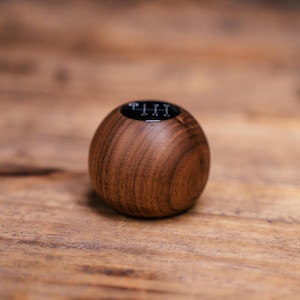 Puede incluir: Un pomo de palanca de cambios esf&eacute;rico de madera con un acabado de grano marr&oacute;n pulido y suave. La parte superior del pomo tiene una tapa circular negra incrustada que muestra un patr&oacute;n de transmisi&oacute;n manual de seis velocidades con los n&uacute;meros 1, 2, 3, 4, 5, 6 y R.