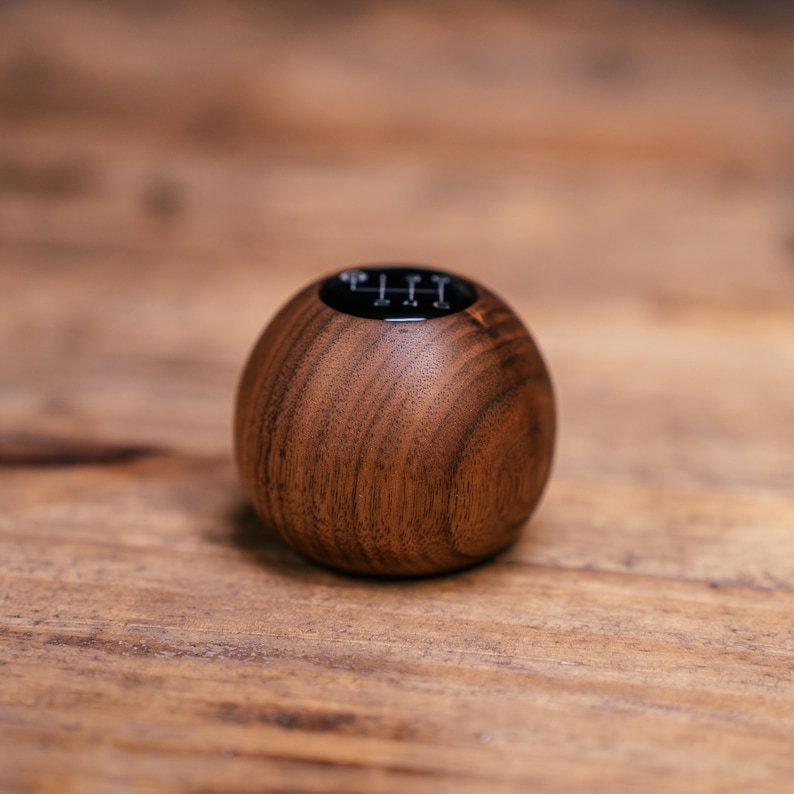 Puede incluir: Un pomo de palanca de cambios esf&eacute;rico de madera con un acabado de grano marr&oacute;n pulido y suave. La parte superior del pomo tiene una tapa circular negra incrustada que muestra un patr&oacute;n de transmisi&oacute;n manual de seis velocidades con los n&uacute;meros 1, 2, 3, 4, 5, 6 y R.