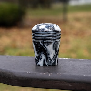 May include: A black and white marbled decorative object with a rounded top and a cylindrical body. The swirling pattern creates a unique visual texture. The object is sitting on a dark surface.