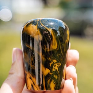 May include: A close-up of a decorative, cylindrical object with a glossy finish. The object features a swirling pattern of dark green and golden yellow colors. The design is reminiscent of marble or a natural stone.