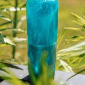May include: A cylindrical, teal-colored object with a marbled effect. The top is rounded, and the base has a ridged design. The object is set against a blurred background of green foliage.