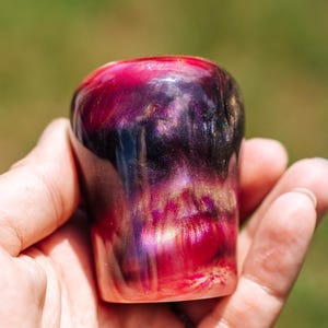 Op de afbeelding: Een handgemaakt hars object met een afgeronde vorm. De kleuren lopen over van diep rood aan de basis naar levendig roze en paars aan de bovenkant, met gouden spikkels. Het object wordt in een hand gehouden.