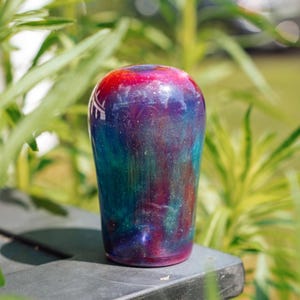 May include: A colorful, cylindrical object with a glossy finish. The top is red, transitioning to purple, blue, and green hues. The object is set against a blurred green background, suggesting an outdoor setting. The design resembles a galaxy.