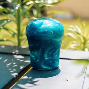 May include: A teal and blue, pearlescent, resin shift knob. The knob has a rounded top and a cylindrical base. The surface has a swirling pattern with flecks of white. The background is blurred, with green foliage and sunlight.
