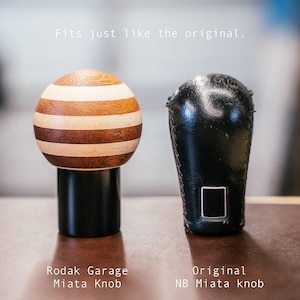 Peut inclure: Un pommeau de levier de vitesse sphérique avec des rayures horizontales alternées en bois clair et foncé sur une base cylindrique noire, à côté d'un pommeau en cuir noir. Texte : Fits just like the original. Rodak Garage Miata Knob. Original NB Miata knob.