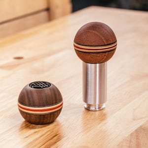 May include: Two wooden shift knobs with a round design. One is attached to a silver metal base. Both knobs are brown with a red and tan stripe. The other knob has a black gear shift pattern on top.