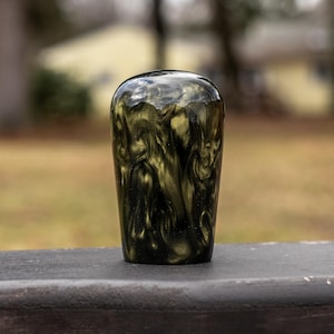 Op de afbeelding: Een glanzend, cilindrisch object met een wervelend patroon van zwart en goud. De bovenkant is afgerond en de basis is plat. Het object staat tegen een vage buitenachtergrond.