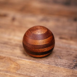 Puede incluir: Un objeto esf&eacute;rico elaborado con m&uacute;ltiples bandas horizontales de madera en diversos tonos de marr&oacute;n, desde el bronceado claro hasta el chocolate oscuro. La superficie muestra un acabado liso y pulido que resalta las vetas naturales.