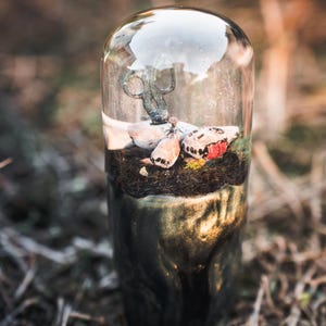 May include: A decorative glass terrarium with a dark, marbled base. Inside, a miniature scene features a small, weathered metal object, a fabric element, and a red accent, set against a backdrop of moss and soil.