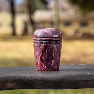 May include: A decorative, cylindrical bottle stopper with a swirling pattern of red, white, and black. The stopper has a rounded top and a ridged body. The product is sitting on a dark wooden surface.