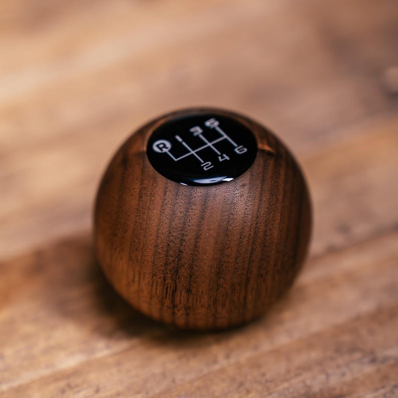 Puede incluir: Un pomo de palanca de cambios esf&eacute;rico de madera marr&oacute;n oscuro con una textura de grano suave. La parte superior del pomo tiene una incrustaci&oacute;n negra circular que muestra un patr&oacute;n de transmisi&oacute;n manual de seis velocidades con R, 1, 2, 3, 4, 5 y 6.
