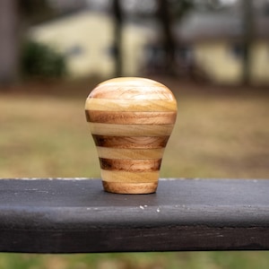 May include: A layered wooden shift knob with a rounded top and flat base. The knob is made from alternating bands of light and dark wood, creating a striped effect. This handcrafted item is designed for a vehicle's gear shift and is a decorative accessory.