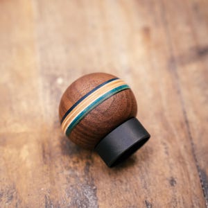 May include: A spherical wooden shift knob, dark brown in colour, with a band of blue, green, and tan stripes. It has a black base and sits on a wooden surface.
