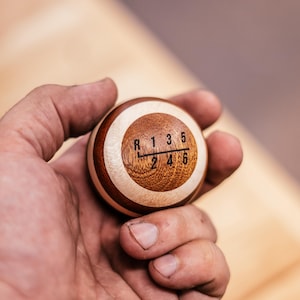 Puede incluir: Un pomo de palanca de cambios esférico hecho de segmentos de madera clara y oscura alternados. La superficie superior presenta un patrón de transmisión manual de seis velocidades grabado en negro con R, 1, 3, 5 arriba y 2, 4, 6 abajo.