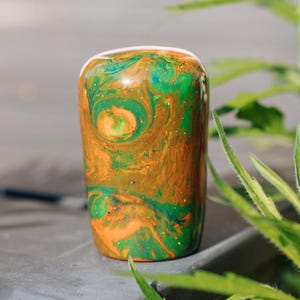 May include: A cylindrical, hand-painted art piece with a swirling pattern of orange, green, and blue. The colors blend together, creating a marbled effect. The piece is displayed outdoors, with green foliage in the background.