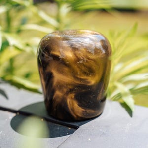 May include: A decorative, cylindrical object with a swirling pattern of gold and black. The glossy surface reflects light, creating a shimmering effect. The object is set against a blurred background of green foliage.