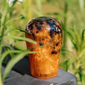 Könnte beinhalten: Ein handgefertigter Schaltknauf aus Kunstharz mit einem einzigartigen Design. Der Knauf hat goldene und bernsteinfarbene Farbtöne mit dunklen Sprenkeln und Glitzer, was einen schimmernden Effekt erzeugt. Das Objekt ist zylindrisch.