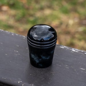 May include: A glossy, black decorative knob with a rounded top and a cylindrical base. The base features white horizontal stripes and a marbled effect. The knob is sitting on a dark gray surface.