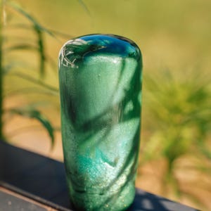 May include: A cylindrical, green decorative object with a rounded top. The object has a shimmering, glittery finish with darker green and teal accents. The background is blurred, with green foliage and a dark surface.