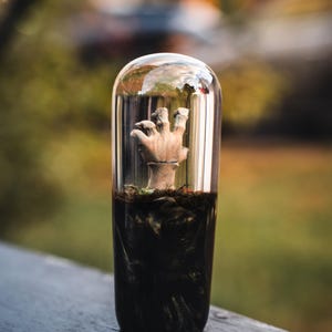 Könnte beinhalten: Ein klares, zylindrisches Glas-Terrarium mit einer modellierten Hand, die aus dunkler Erde ragt. Die Hand hat eine hellbraune Farbe, und die Erde ist tiefschwarz. Das Terrarium steht vor einem verschwommenen Hintergrund im Freien.