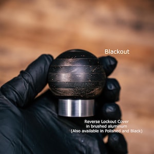 Puede incluir: Un pomo de palanca de cambios esf&eacute;rico con un acabado oscuro y texturizado con bandas horizontales. La base es una cubierta cil&iacute;ndrica de bloqueo de marcha atr&aacute;s de aluminio cepillado. Texto: Blackout y Reverse Lockout Cover in brushed aluminum.