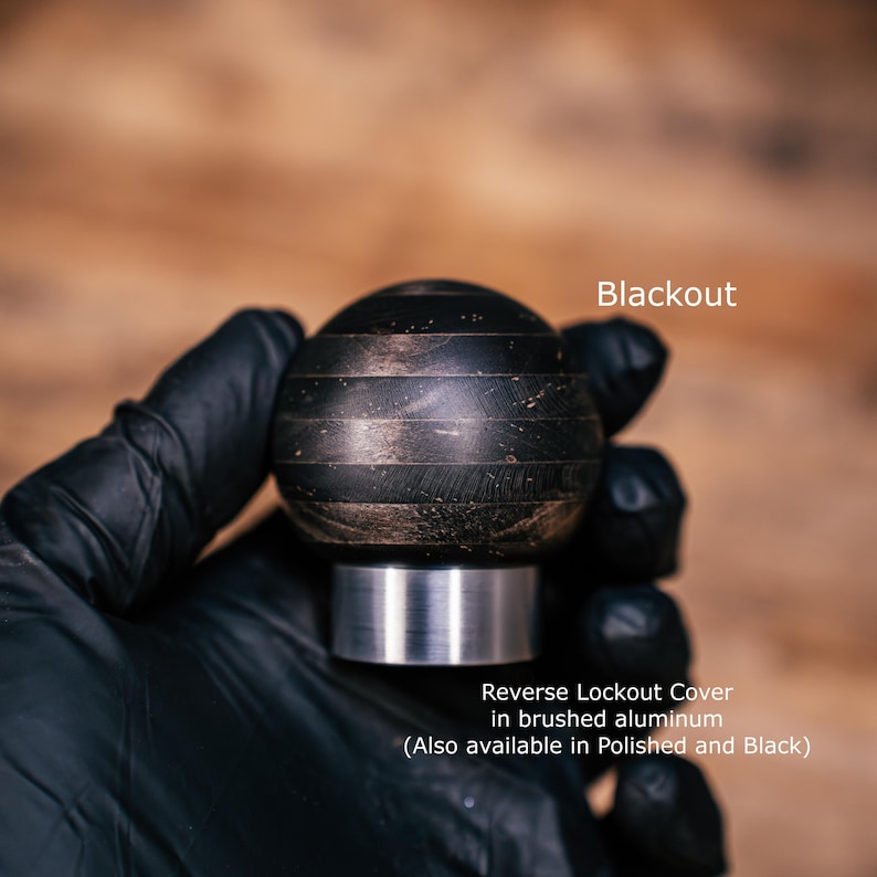 Puede incluir: Un pomo de palanca de cambios esf&eacute;rico con un acabado oscuro y texturizado con bandas horizontales. La base es una cubierta cil&iacute;ndrica de bloqueo de marcha atr&aacute;s de aluminio cepillado. Texto: Blackout y Reverse Lockout Cover in brushed aluminum.