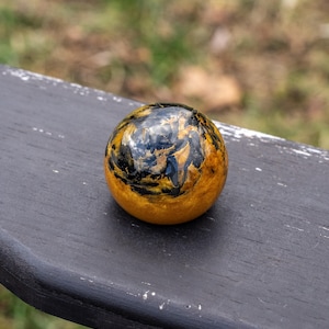 May include: A decorative sphere with a polished, glossy finish. The orb features a blend of gold, black, and brown tones, creating a marbled effect. The sphere is resting on a dark, weathered surface, with a blurred natural background.