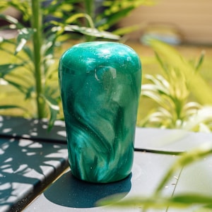 May include: A cylindrical, emerald green object with a glossy finish. The surface has a swirling, marbled effect, with lighter green and silver accents. The object is set against a blurred background of green foliage and a light-coloured building.