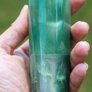 May include: A cylindrical, green, decorative object with a metallic sheen. The object is held in a hand and has a smooth, polished surface. The color transitions from a lighter green at the top to a darker shade at the base. The object is approximately 4 inches tall.