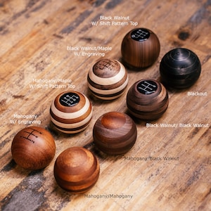 Puede incluir: Una colección de pomos de cambio de madera en varios estilos y acabados. Los pomos incluyen opciones como Black Walnut, caoba y Blackout, algunos con patrones de cambio o grabados. La imagen muestra las diferentes vetas y diseños de la madera.