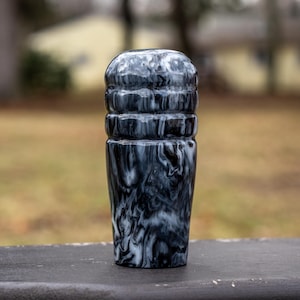 May include: A black and white marbled resin shift knob. The shift knob has a cylindrical shape with a rounded top and a series of horizontal ridges. The swirling pattern creates a unique, decorative appearance. The object is set against a blurred outdoor background.