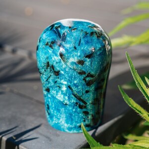 Könnte beinhalten: Eine blau-schwarze dekorative Vase mit glänzendem Finish. Die Vase hat eine abgerundete Oberseite und eine strukturierte Oberfläche. Die Vase steht auf einer dunkelgrauen Oberfläche, mit grünen Blättern im Hintergrund.