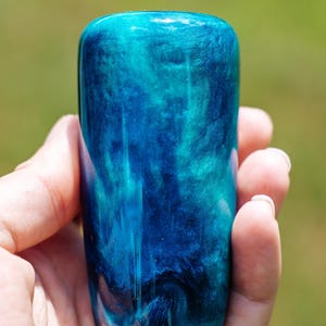 May include: A hand holding a cylindrical object with a rounded top. The object is a mix of blues and teals, with a swirling, marbled effect. The colours blend together, creating a unique design. The object appears to be made of resin, with a glossy finish.
