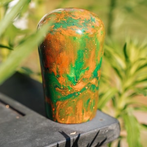 Könnte beinhalten: Ein zylindrisches Objekt mit abgerundeter Oberseite, das einen lebendigen Wirbel aus orangefarbenen und grünen Farben aufweist. Das Objekt befindet sich vor einem verschwommenen Hintergrund aus grünem Laub und ruht auf einer dunklen Oberfläche.