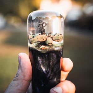 May include: A clear, cylindrical resin object with a dark, glittery base and a miniature scene inside. The scene includes a small, detailed sculpture and a textured, green element. The top is rounded, and the object is held in a hand.