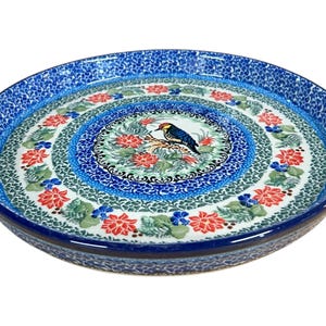 Puede incluir: Un plato de cerámica redondo con un diseño floral azul y blanco. El centro presenta un pájaro posado en una rama, rodeado de flores rojas y azules. El borde tiene un patrón azul texturizado.