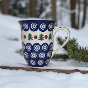 May include: A tall ceramic mug with a blue and white pattern featuring green and red circles and small green Christmas trees. The mug has a white handle and is set against a snowy background with evergreen branches.