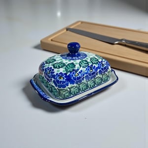 May include: A ceramic butter dish with a blue floral pattern and a matching lid. The dish is rectangular with a slightly curved base and a small knob on top of the lid. A wooden cutting board and knife are in the background.