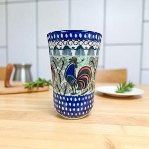 May include: A tall ceramic tumbler with a blue and white rooster design. The tumbler features a blue rim and base with white polka dots. The body of the tumbler has a rooster illustration with red, blue, and black accents, surrounded by a leaf pattern.