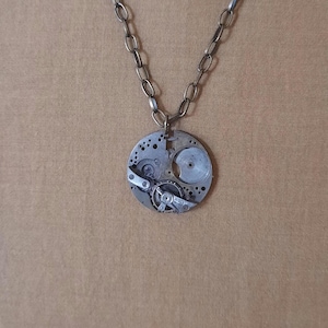 May include: A silver necklace with a round pendant featuring a circular clockwork design. The pendant has exposed gears and a silver chain.