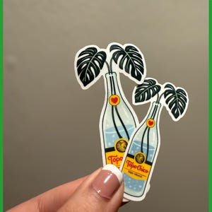 May include: Two stickers featuring a Topo Chico bottle design with a monstera leaf arrangement. The bottles are clear with blue liquid and a yellow label. Each bottle has a red heart detail. The stickers are held by a person.